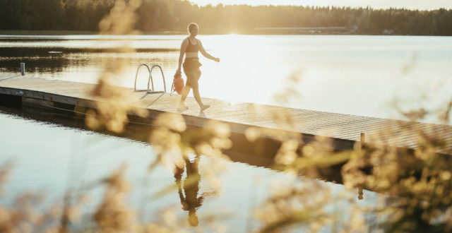 Nainen kävelee märkäpuvussa laiturilla kesäisessä järvimaisemassa.