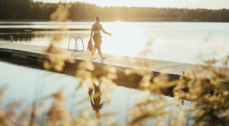 Nainen kävelee märkäpuvussa laiturilla kesäisessä järvimaisemassa.