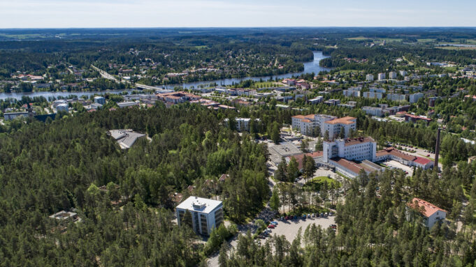 Ilmakuva Sairaalanmäestä ja Kuusankoskesta. Valkoisia sairaalarakennuksia, taloja metsän ympäröimänä ja taustalla Kymijoki.