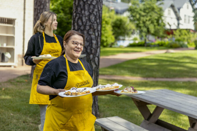 Kaksi henkilöä keltaisissa essuissa kantaa ruoka-annoksia pöytään ulkona.