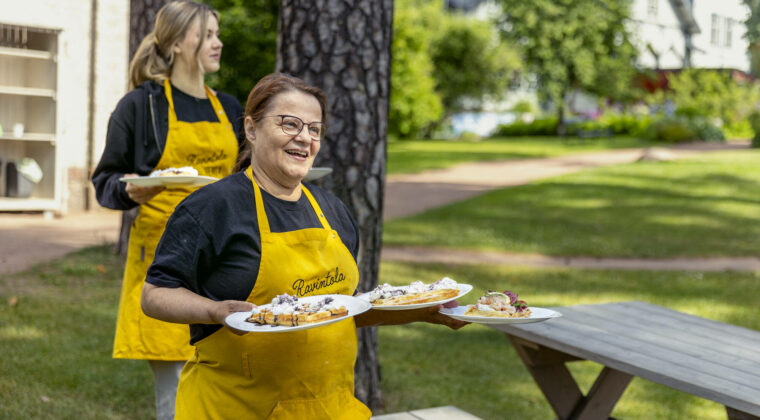 Kaksi henkilöä keltaisissa essuissa kantaa ruoka-annoksia pöytään ulkona.